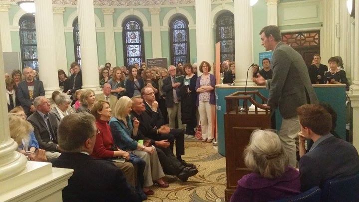 Picture of poet Martin Dyar at the podium reading from his work to a crowd of people in a room with columns. Paula Cunningham, too, reads from her work at this event