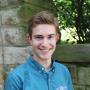 Bailey Bradford, wearing a chambray shirt, smiling, and standing in front of a stone wall