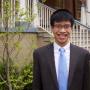 Caleb Yip, wearing a black suit and light blue tie, standing in front of some stairs, smiling