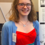 Jane, wearing a red dress with a light blue sweater, smiling in front of an academic conference display