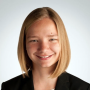 Katherine Schmidt, wearing a black blazer, smiling and standing in front a grey wall