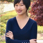 Rebecca, wearing a blue sweater and red lipstick, smiling with her arms crossed, standing in front of a scenic area