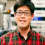 Matthew, wearing a red and green plaid shirt and black glasses, standing in a lab