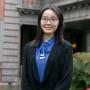 Zezhou, wearing a blue blouse, silver necklace, and black sweater, standing in front of a grey stone building