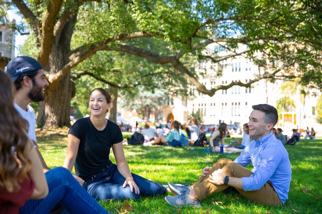 A small group of students sitting on the grass in a circle smiling.