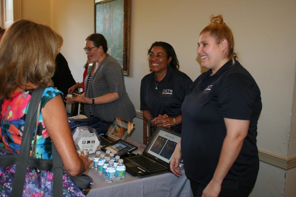 CETS staff standing behind a table at New Faculty Orientation in 2016 with form, a presentation, and free goodies talking to a faculty member about CETS