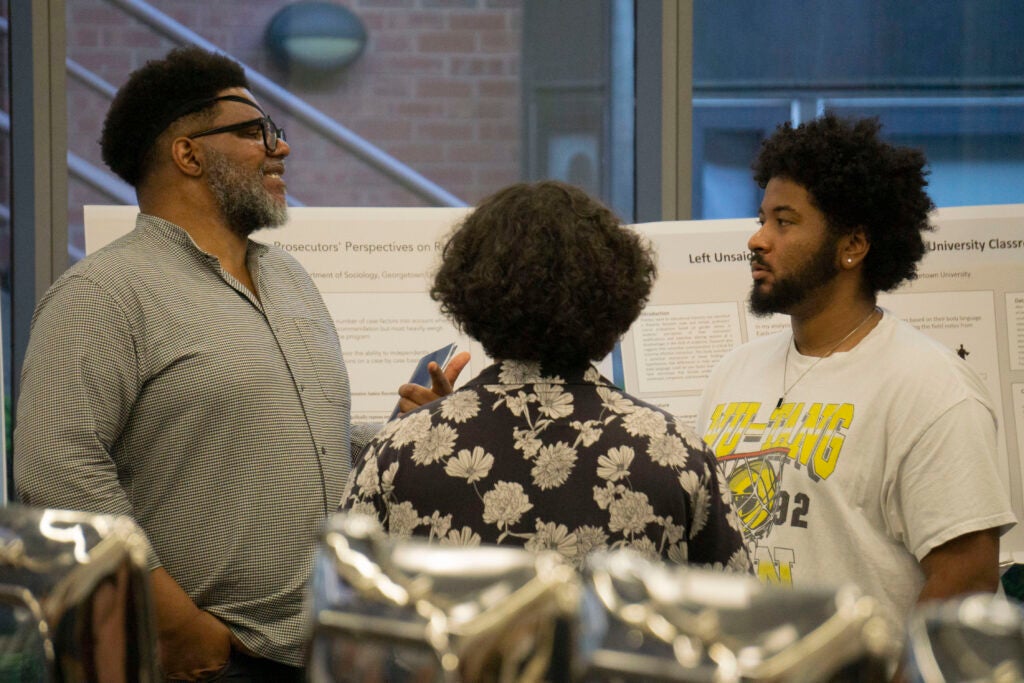 Professor Corey Fields talks with seniors Eric Perez and Langston Lee while standing in front of several presentations