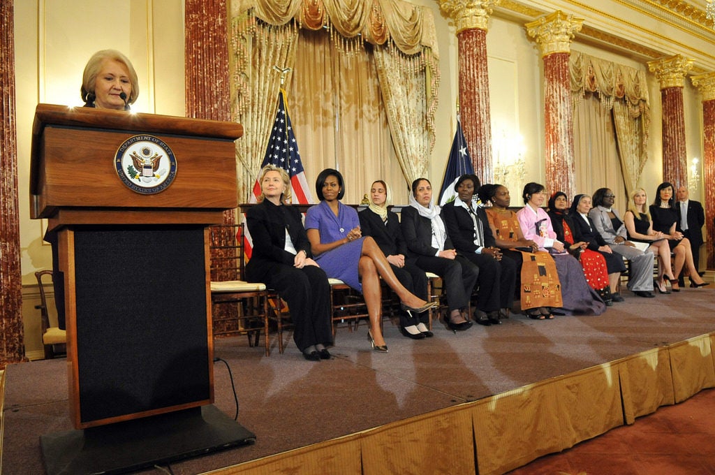 Ambassador Melanne Verveer talking at a podium in front of a panel of women including Hilary Clinton and Michelle Obama